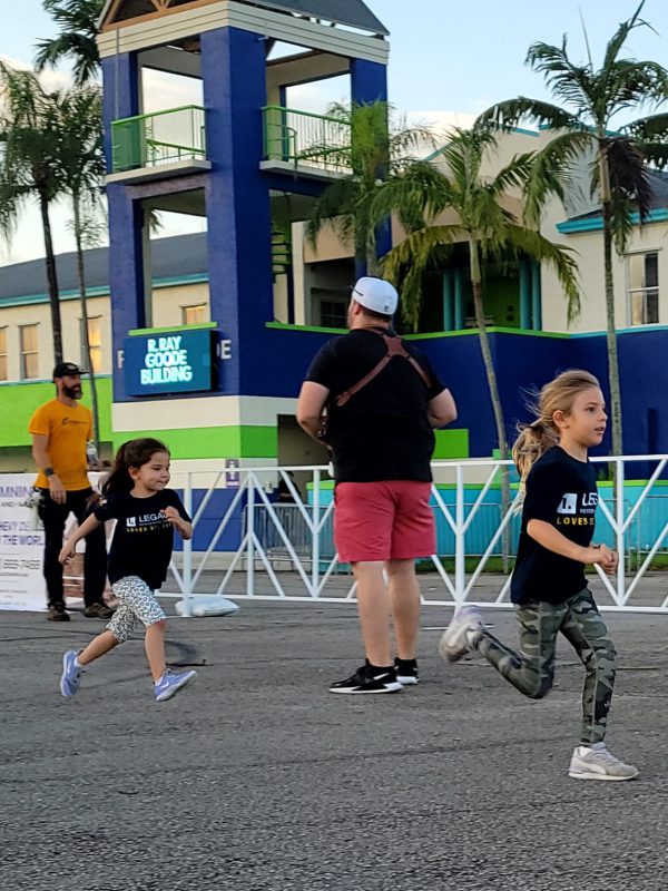 Two young girls wearing Legacy Residential Group shirts running at the 2023 St. Jude Walk/Run event, with adults and a colorful building in the background.