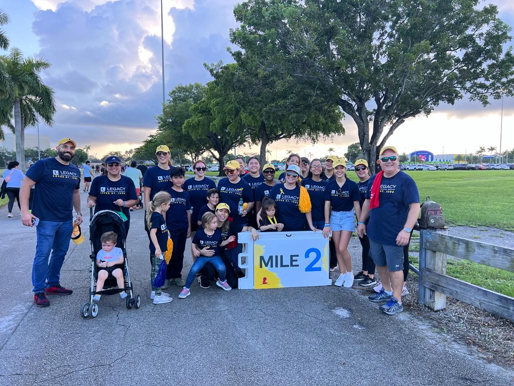 A group of participants from Legacy Residential Group posing together at the 2023 St. Jude Walk/Run event, standing around a Mile 2 marker with trees and a cloudy sky in the background.